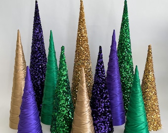 May include: A collection of cone-shaped Christmas trees decorated with sequins in purple, green, and gold. The trees are made of fabric and are arranged in a row on a white surface.