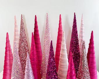 May include: A collection of pink and fuchsia Christmas tree cones made of fabric and sequins. The cones are arranged in a semi-circle on a white surface.
