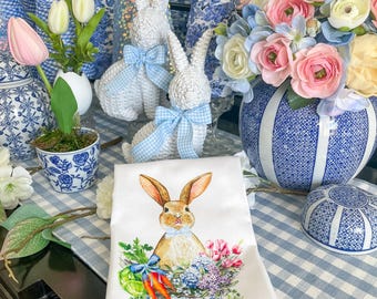 May include: A white kitchen towel with a blue and white floral design and a brown bunny rabbit holding a basket of carrots and flowers. The towel is on a blue and white gingham tablecloth.