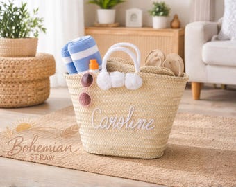 Könnte beinhalten: Eine geflochtene Stroh-Tragetasche mit weißen Griffen und Bommeln, gefüllt mit einem blau-weiß gestreiften Handtuch, Sonnenbrille und Sandalen. Die Tasche ist mit dem Namen "Caroline" in weißer Schrift personalisiert.