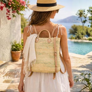 May include: A straw backpack with tan leather straps and a handle. The bag is rectangular with a buckle closure. A white scarf is draped over the top. The text "Bohemian Straw" is visible. The background includes a pool and flowers.