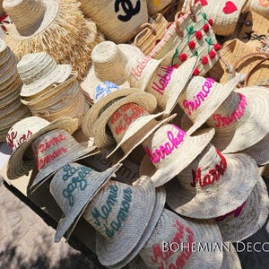 May include: A collection of straw hats with embroidered lettering in various colors, including pink, blue, and green. The hats are arranged in a pile, with some of them overlapping. The hats are all made of natural straw and have a wide brim.