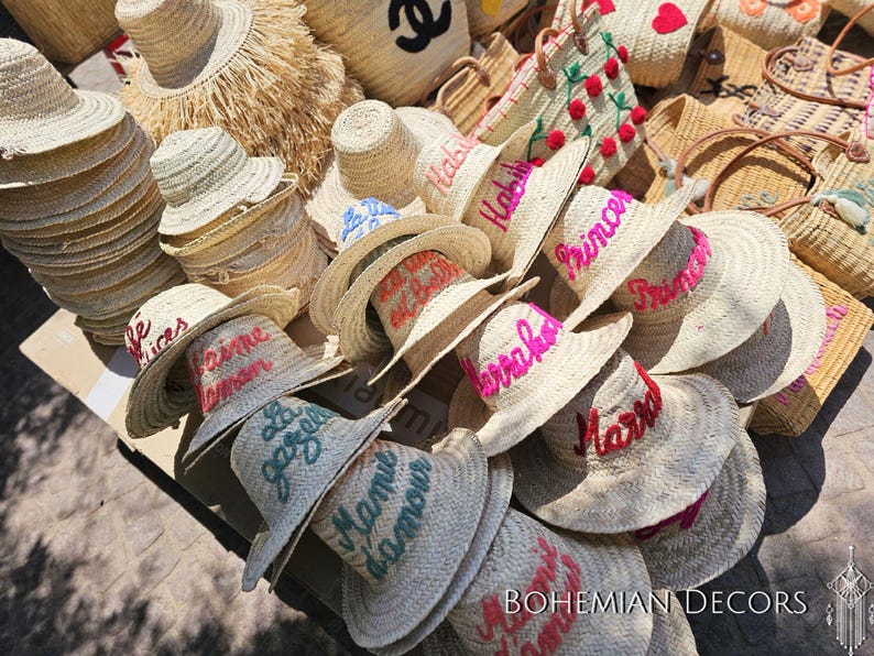 May include: A collection of straw hats with embroidered lettering in various colors, including pink, blue, and green. The hats are arranged in a pile, with some of them overlapping. The hats are all made of natural straw and have a wide brim.