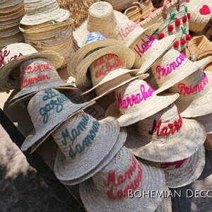 May include: A collection of straw hats with various embroidered phrases in different colors, including "Habibi", "Marrakech", "Mama", and "Le Jardin".