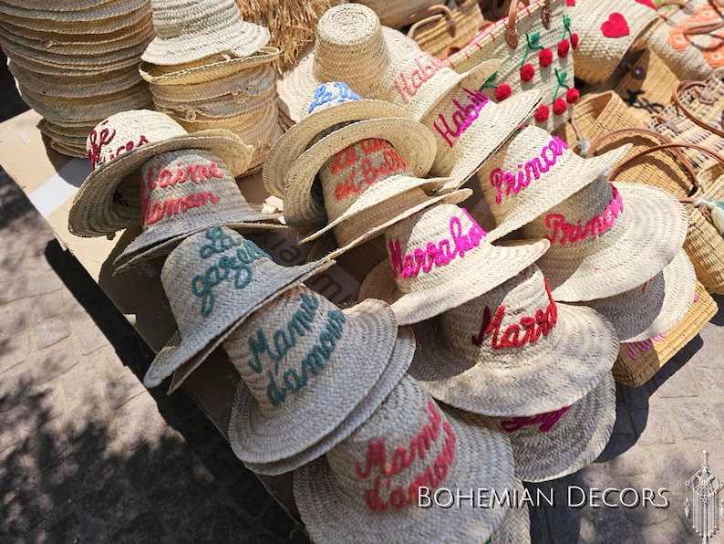 May include: A collection of straw hats with various embroidered phrases in different colors, including "Habibi", "Marrakech", "Mama", and "Le Jardin".