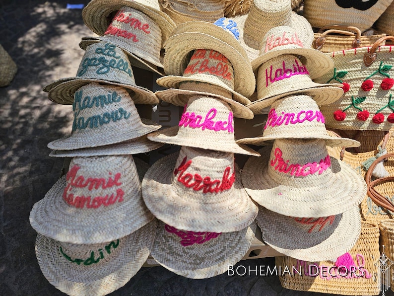 May include: A collection of straw hats with colorful embroidered lettering. The hats are arranged in a stack, with the top hat showing the text "Habibi".