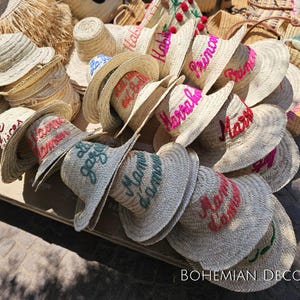 May include: A pile of straw hats with various embroidered phrases in pink, green, and blue. The phrases include "Habibi", "Princesa", "Marrakech", and "Mon Amour".
