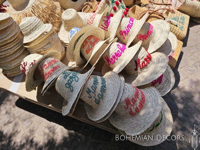 May include: A pile of straw hats with various embroidered phrases in pink, green, and blue. The phrases include "Habibi", "Princesa", "Marrakech", and "Mon Amour".