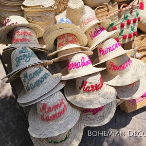 May include: A collection of straw hats with colorful embroidered lettering. The hats are stacked on top of each other and feature various phrases like "Habib", "Marrakech", and "Mamie d'amour".