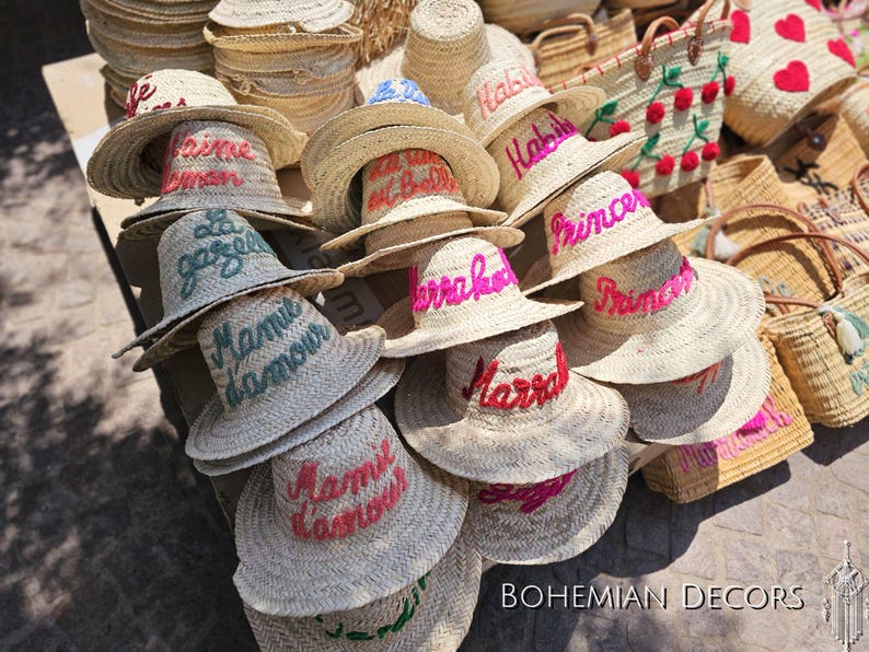 May include: A collection of straw hats with colorful embroidered lettering. The hats are stacked on top of each other and feature various phrases like "Habib", "Marrakech", and "Mamie d'amour".