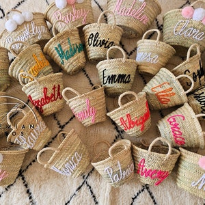 K&ouml;nnte beinhalten: Eine Sammlung kleiner Strohtaschen mit personalisierten Namen, die in verschiedenen Farben gestickt sind. Die Taschen sind auf einem gemusterten Teppich angeordnet.
