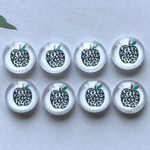 May include: Eight clear glass magnets with a white background. Each magnet features a black and white leopard print apple design with a green stem and leaf. The magnets are arranged in two rows of four.