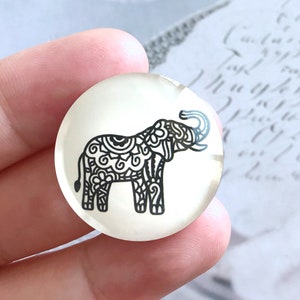 May include: A clear, round glass cabochon featuring a black, ornate elephant design. The elephant is detailed with swirling patterns and has its trunk raised. The cabochon is held between fingers, with a blurred background.