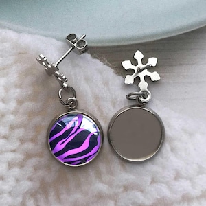 May include: A pair of silver-colored stud earrings. One earring features a round, glass-covered design with a black and purple zebra stripe pattern. The other earring has a blank, round, silver-colored surface. The earrings have snowflake-shaped accents.