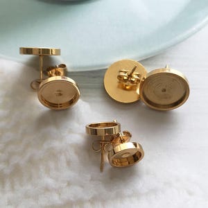 May include: Gold-coloured earring posts with flat, round settings. The earring posts have a post and a flat disc on the back for securing the earring. The settings are designed to hold a decorative element, such as a cabochon or a resin design.