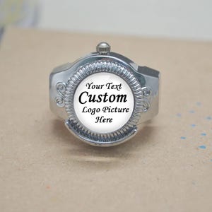 May include: A silver-toned ring with a circular face, featuring the text "Your Text Custom Logo Picture Here" in black. The ring has a decorative design around the face and a small knob on top. The ring is set against a neutral background.