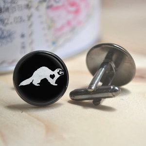 May include: A pair of black cufflinks featuring a white silhouette of a ferret with a heart on its chest. The cufflinks are displayed on a wooden surface.