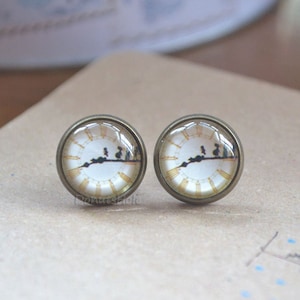 May include: A pair of bronze-colored earrings with a clock face design. The clock face is white with black Roman numerals and black hands. The clock face is surrounded by a gold-colored ring.