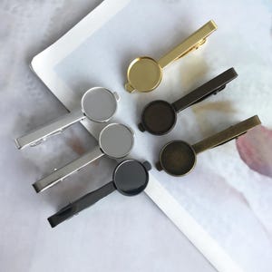 Puede incluir: Una variedad de pasadores de corbata metálicos en dorado, plateado, negro y bronce. Cada pasador presenta una barra rectangular con un engaste redondo, perfecto para añadir un diseño personalizado. Los pasadores están dispuestos sobre una superficie blanca y gris.