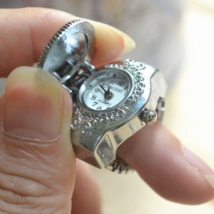 May include: A silver-toned ring watch with a hinged cover, featuring a white clock face with black numerals and hands. The ring has an ornate, decorative design around the watch face. The watch is being held up by a finger.