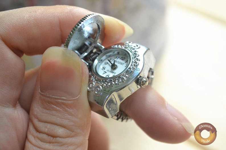 May include: A silver-toned ring watch with a hinged cover, featuring a white clock face with black numerals and hands. The ring has an ornate, decorative design around the watch face. The watch is being held up by a finger.