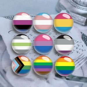 Puede incluir: Nueve cabujones de vidrio circulares dispuestos en una cuadr&iacute;cula, cada uno con un dise&ntilde;o de bandera de orgullo diferente. Las banderas incluyen los dise&ntilde;os genderqueer, transg&eacute;nero, pansexual, ag&eacute;nero, bisexual, asexual, progress pride y dos variantes del arco&iacute;ris.