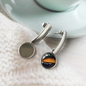 May include: Silver-coloured earrings. Each earring has a curved bar and a round pendant. One pendant is a solid grey colour, while the other features a colourful sunset design with orange, yellow, and blue hues.