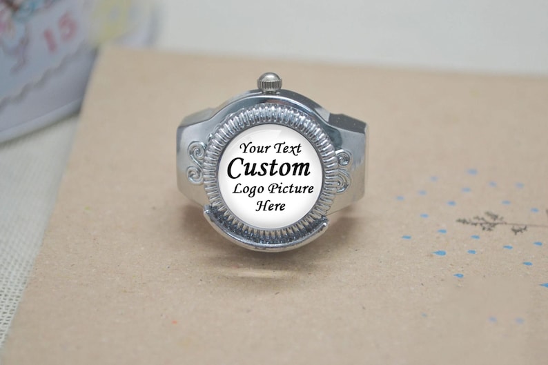 May include: A silver-toned ring with a circular face, featuring the text "Your Text Custom Logo Picture Here" in black. The ring has a decorative design around the face and a small knob on top. The ring is set against a neutral background.