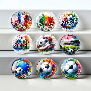 Puede incluir: Nueve cabujones redondos de vidrio con diseños de fútbol. Los diseños incluyen balones de fútbol, banderas, botas de fútbol y la palabra "Soccer". Los cabujones están rodeados de motivos florales y una escena de estadio.