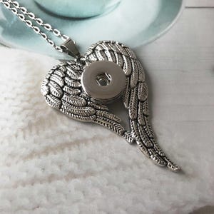 May include: Silver-toned necklace featuring a pendant shaped like angel wings. The wings are detailed with feather textures and surround a circular snap button. The necklace chain is also silver and delicate.