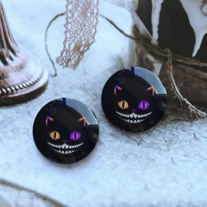 Può includere: Due cabochon rotondi in vetro nero con un gatto nero sorridente con occhi viola e arancioni. La bocca del gatto è aperta e mostra denti affilati.