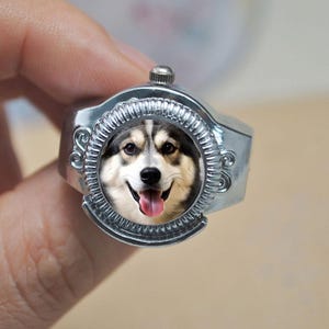 May include: Silver-toned ring featuring a smiling dog portrait. The ring has an ornate, circular setting around the dog's face, with a small knob on top. The dog has black, white, and brown fur, with its tongue sticking out.