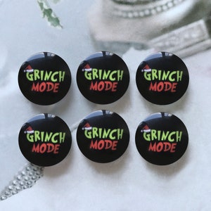 以下が含まれることがあります： 「GRINCH MODE」の文字が緑と赤で書かれた、サンタ帽が「GRINCH」の上に描かれた、黒い丸いボタンが6つ。ボタンは光沢があり、わずかにドーム型になっています。
