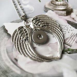 May include: A silver-colored necklace with a pendant shaped like angel wings. The wings encircle a circular snap button. The necklace chain is also silver. The background is a soft, blurred image of a floral arrangement and handwritten script.