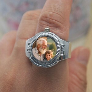 May include: A silver ring featuring a circular photo of an older man with a white beard and a young boy. The ring has a decorative, textured bezel around the photo and a wide band. The ring is worn on a finger.
