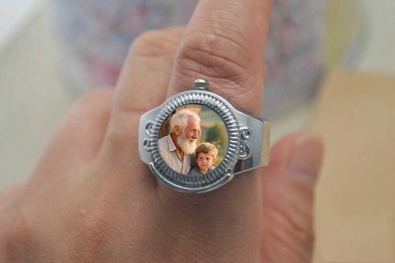 May include: A silver ring featuring a circular photo of an older man with a white beard and a young boy. The ring has a decorative, textured bezel around the photo and a wide band. The ring is worn on a finger.