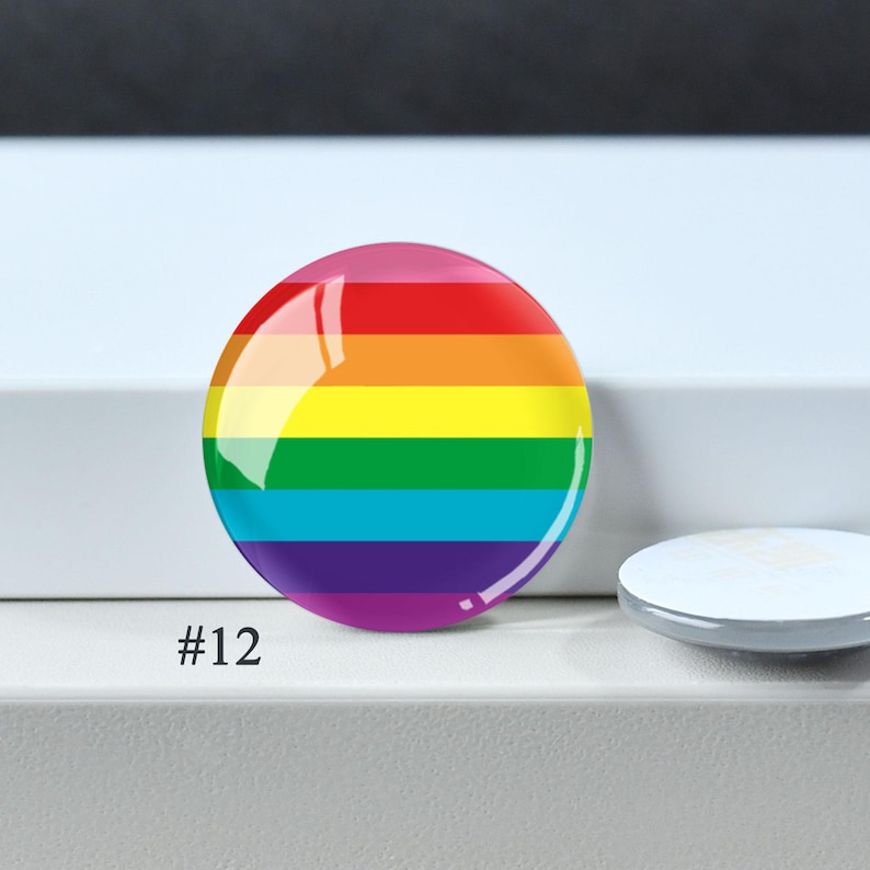 Puede incluir: Un bot&oacute;n circular con un dise&ntilde;o de bandera arco&iacute;ris horizontal con franjas rojas, naranjas, amarillas, verdes, azules y moradas. El bot&oacute;n tiene un acabado brillante y est&aacute; etiquetado con el n&uacute;mero 12 en la esquina inferior izquierda.