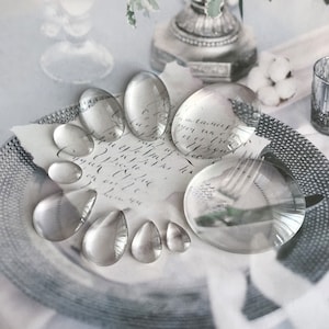 May include: A collection of clear glass magnifying lenses of various sizes and shapes, arranged on a white surface with a handwritten note.