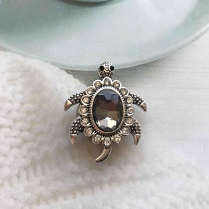 以下が含まれることがあります： 中央に大きな濃いグレーのラインストーンが付いた、銀色のカメのブローチ。カメは、より小さなクリアなラインストーンで囲まれています。