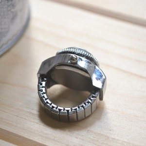 May include: A silver-toned ring watch with a round face and a stretch band. The watch has a detailed bezel and a classic design. The ring watch is displayed on a light wooden surface.