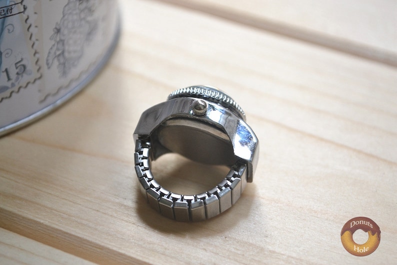 May include: A silver-toned ring watch with a round face and a stretch band. The watch has a detailed bezel and a classic design. The ring watch is displayed on a light wooden surface.