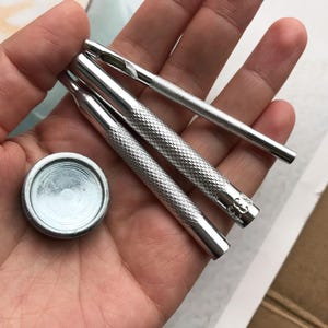 Könnte beinhalten: Drei silberne Metallwerkzeuge zum Stanzen von Löchern in Leder, mit einem runden silbernen Metallstück zum Befestigen an Leder.