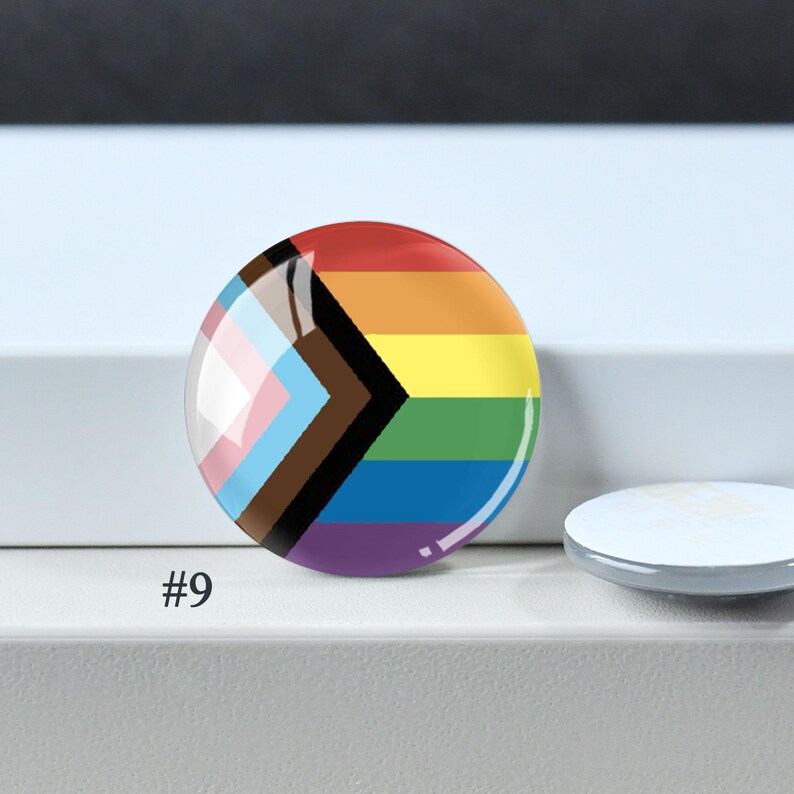 Puede incluir: Una chapa redonda con el dise&ntilde;o de la bandera Progress Pride, que presenta franjas horizontales de color rojo, naranja, amarillo, verde, azul y morado, junto a un patr&oacute;n de chevr&oacute;n en negro, marr&oacute;n, azul claro, rosa y blanco. Texto #9 visible.