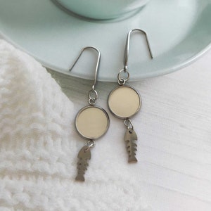 May include: A pair of silver dangle earrings with a round, blank, silver charm and a fishbone charm hanging from each earring.