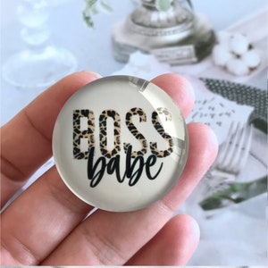 Może przedstawiać: Przezroczysty, okrągły szklany przycisk do papieru z napisem "BOSS babe". Słowo "BOSS" jest w wzorze w cętki, a "babe" jest napisane czarną czcionką. Przycisk do papieru jest trzymany w dłoni.