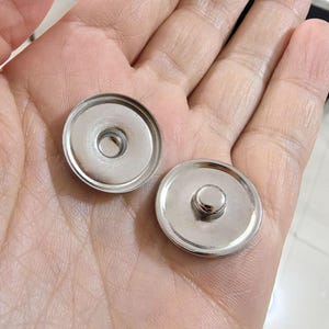 Può includere: Due bottoni a pressione lucidi color argento. Un bottone a pressione ha un foro centrale e l'altro ha un bottone rialzato. Le chiusure sono rotonde e sembrano essere in metallo. Sono mostrate su uno sfondo neutro.