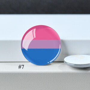 Puede incluir: Una chapa redonda con un dise&ntilde;o tricolor horizontal que presenta una franja superior de color rosa brillante, una franja central de color lavanda y una franja inferior de color azul real. La superficie es brillante. El n&uacute;mero #7 est&aacute; impreso cerca.
