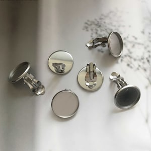 May include: Five silver-toned earring settings with round glass or metal insets. The settings have a clip-on design.