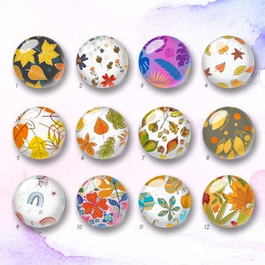 May include: Twelve round glass cabochons with diverse, colourful designs. The designs feature leaves, flowers, and abstract patterns in orange, yellow, blue, and green. Each cabochon has a glossy finish and is set against a white background.