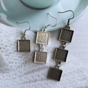 May include: Three pairs of square-shaped earrings with textured surfaces. One pair is a single square, another has two squares, and the third has three squares. The earrings have silver and bronze-toned metal hooks and frames, displayed on a white, textured surface.